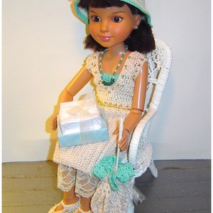 May include: A doll wearing a crocheted outfit, hat, and shoes, seated in a white wicker chair. The doll holds a gift box and a crocheted bag. The outfit is cream-colored with gold accents and turquoise details. The hat has a white lace bow.