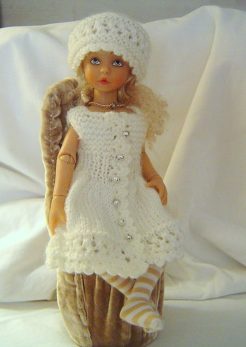 May include: A white porcelain doll wearing a white crocheted dress and hat. The doll is sitting on a brown velvet chair.