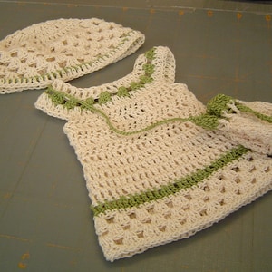 Op de afbeelding: Een gehaakt baby-outfit in wit en groen. De outfit bestaat uit een jurkje en een bijpassende muts.