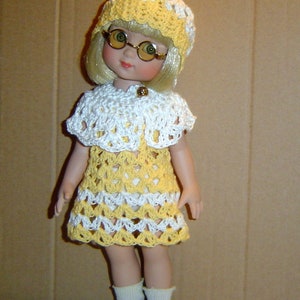 Peut inclure: Une poupée vêtue d'une robe, d'un chapeau et d'une cape en crochet jaune et blanc. La poupée a les cheveux blonds, des lunettes rondes, des chaussettes blanches et des chaussures noires. La robe a un ourlet festonné et une fermeture à bouton sur la cape.