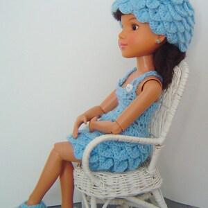 May include: A doll wearing a light blue crocheted dress and hat, sitting on a white wicker chair. The doll has brown hair and is wearing blue and white crocheted shoes.