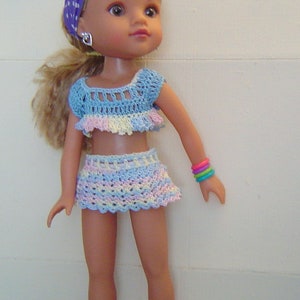 Könnte beinhalten: Eine Puppe, die ein hellblaues und rosafarbenes gehäkeltes Bikini-Oberteil und einen Rock trägt. Die Puppe hat langes blondes Haar und trägt ein lila-weißes gepunktetes Stirnband. Die Puppe trägt rosa Schuhe und einen silbernen Ohrring am linken Ohr.