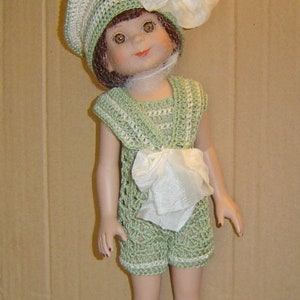 May include: A porcelain doll wearing a crocheted outfit. The doll has brown hair and eyes, and is dressed in a green and white crocheted hat, shorts, and top. A white bow is attached to the hat and the waist of the outfit.