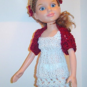 May include: A doll wearing a handmade white crochet dress and a burgundy crochet shrug. The doll has blue eyes, blonde hair, and a headband with burgundy flowers. The dress has a detailed openwork design.