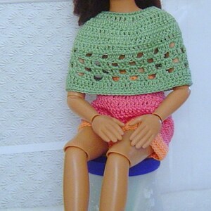 May include: A doll wearing a handmade crochet outfit. The outfit includes a green top, a pink and orange skirt, and green booties with pink trim. The doll also wears a matching hat.