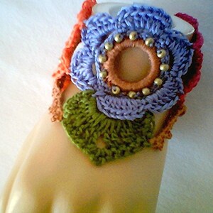 May include: A crocheted bracelet with a purple flower design, green leaves, and orange accents. The flower has a brown center with gold beads around it.