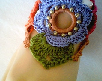 May include: A crocheted bracelet with a purple flower design, green leaves, and orange accents. The flower has a brown center with gold beads around it.