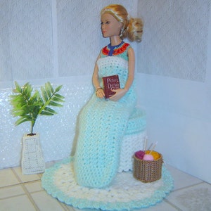 May include: A doll wearing a crocheted dress and seated on a crocheted stool, holding a book titled "Picture of Dorian Gray". A small basket with yarn and knitting needles sits nearby. A small plant in a white vase is also present.