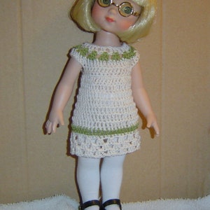 May include: A doll in a cream crochet dress with green detailing, blonde hair, and round glasses. The doll wears white tights and black Mary Jane shoes. The doll is standing on a white surface.