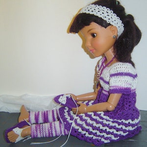 May include: A doll wearing a handmade crochet outfit. The outfit includes a white headband, a white and purple striped dress, matching leg warmers, and sandals. The doll has dark hair and is sitting down.