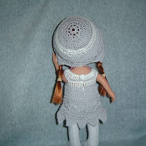 May include: A doll wearing a handmade gray crochet dress and matching hat. The dress has a scalloped hem and white accents. The hat is circular with a white brim. The doll has red hair in two braids.