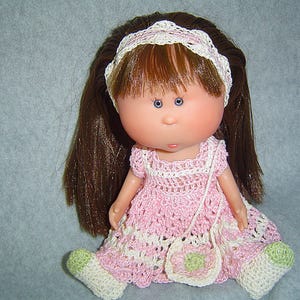 Puede incluir: Muñeca con cabello castaño, ojos azules y un vestido de ganchillo rosa y blanco. La muñeca tiene un bolso y una diadema a juego. Los pies de la muñeca llevan zapatos de ganchillo blancos y verdes.