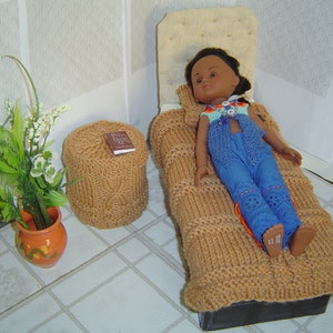 May include: A doll dressed in blue pants and a blue vest with a floral top, resting on a knitted brown blanket. A matching knitted ottoman with a book on top sits nearby. A small orange pot with green plants and white flowers is in the corner.