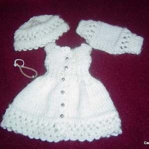 May include: White knitted doll dress with a white knitted hat and matching mittens. The dress has a button closure and a lace trim.