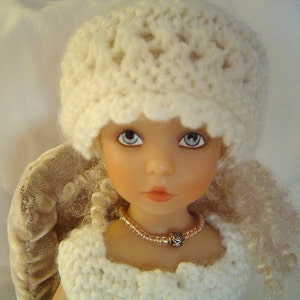 May include: A white crocheted hat with a scalloped edge sits on the head of a doll with blonde curly hair. The doll is wearing a white crocheted dress and a beaded necklace with a rose charm.