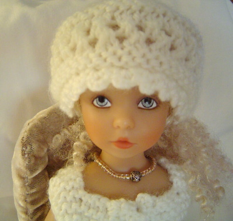 May include: A white crocheted hat with a scalloped edge sits on the head of a doll with blonde curly hair. The doll is wearing a white crocheted dress and a beaded necklace with a rose charm.