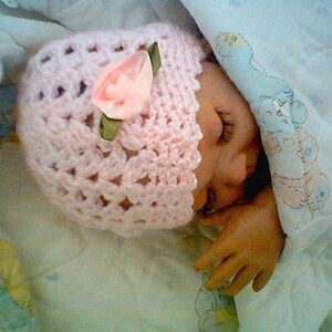 May include: A pink crocheted baby hat with a pink flower embellishment. The hat is on a white blanket with a cartoon print.