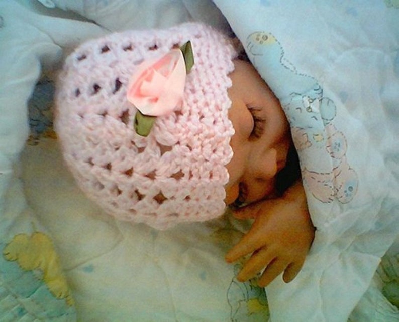May include: A pink crocheted baby hat with a pink flower embellishment. The hat is on a white blanket with a cartoon print.