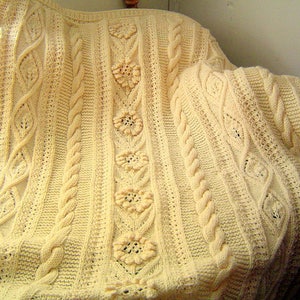 May include: A cream-colored, hand-knitted blanket with a cable knit design and floral accents. The blanket is draped over a chair, showcasing its intricate patterns and textures. The soft, warm blanket is perfect for home decor.