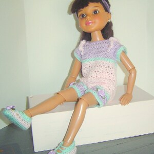 May include: A doll wearing a handmade crochet outfit. The outfit includes a short-sleeved top and shorts in pastel colors: lavender, mint green, and pink. The doll also wears matching crochet shoes with rose accents. The doll has dark hair and a headband.