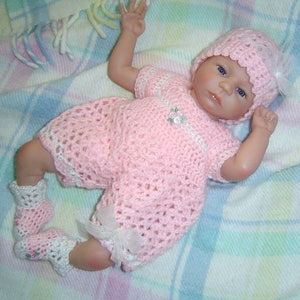 May include: A pink crocheted baby outfit with a white crocheted hat. The outfit has a flower design on the front and a bow on the leg.