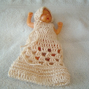 May include: A crocheted baby doll dress with a hood in a cream color. The dress has a lacy pattern and is made with a soft yarn.