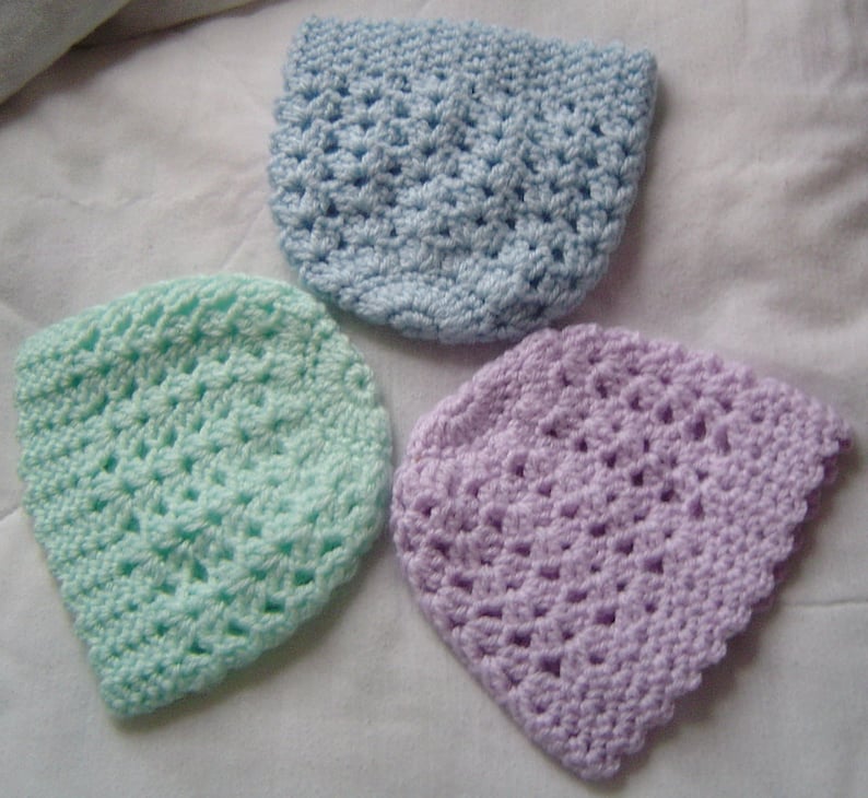 May include: Three crocheted hats in different colors: light blue, light green, and light purple. The hats are made with a simple stitch pattern and have a scalloped edge.