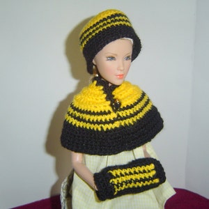 May include: A doll dressed in a handmade yellow and black striped crochet hat, cape, and fingerless mittens. The hat is a cloche style, and the cape fastens with a button. The doll is wearing a yellow and white dress.