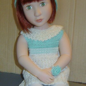 Puede incluir: Una muñeca con el pelo rojo que lleva un vestido de ganchillo blanco y azul claro y un gorro de ganchillo azul claro.