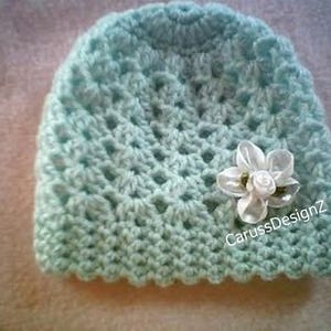 May include: A light blue crocheted baby hat with a white flower accent. The hat has a scalloped edge and the text "CarussDesignZ" is visible on the side.