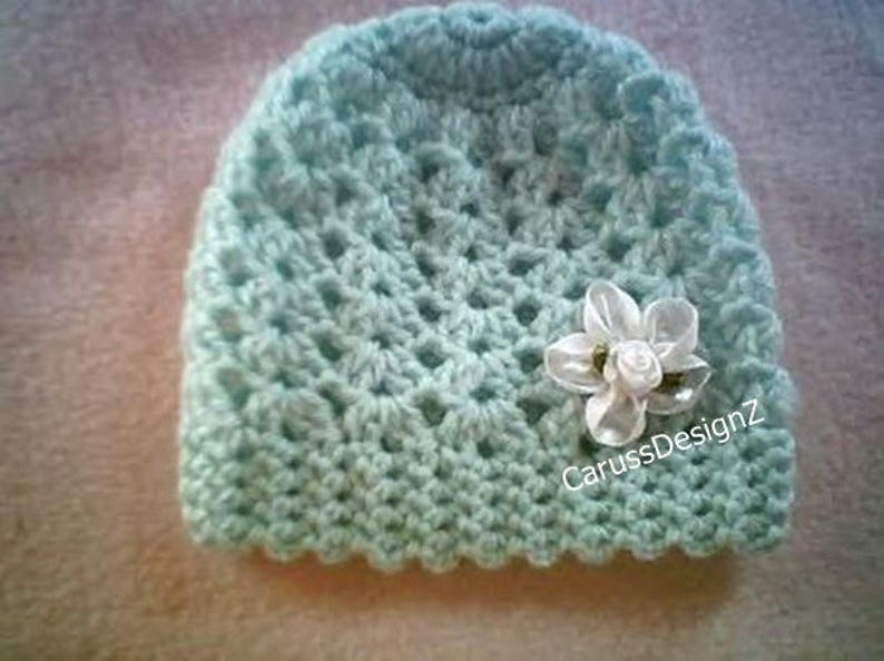 May include: A light blue crocheted baby hat with a white flower accent. The hat has a scalloped edge and the text "CarussDesignZ" is visible on the side.