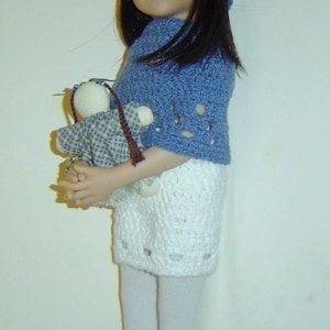 May include: A doll wearing a blue crocheted hat with a flower, a matching shawl, and a white crocheted skirt. The doll is holding a small stuffed animal and wearing blue crocheted booties. The doll has black hair and is wearing white tights.
