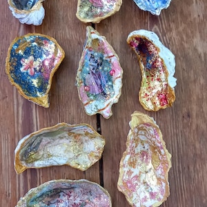 May include: A selection of decorated oyster shells with painted and embellished interiors. The shells display diverse designs, including floral patterns, gold leaf accents, and images of figures. The shells are arranged on a wooden surface.