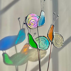 May include: Three stained glass snail ornaments on metal stakes. One snail has an iridescent purple shell and blue body, one has a green body and light blue shell, and one has an orange body with a clear textured shell. Each has two wire antennae.