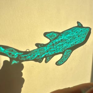 May include: A teal-colored, stylized shark silhouette with intricate line details and small dot patterns. The shark's outline is sharply defined, and it appears to be a handmade craft or art piece. The image is set against a light background.