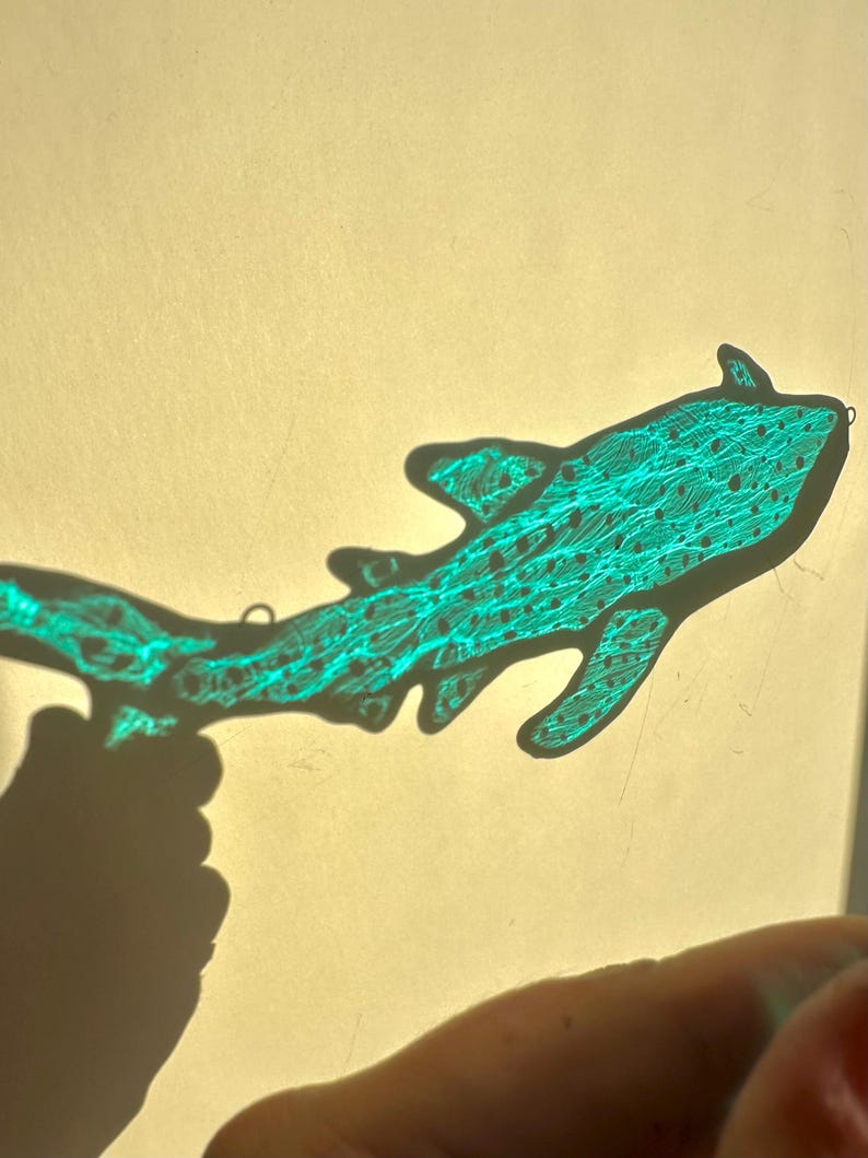 May include: A teal-colored, stylized shark silhouette with intricate line details and small dot patterns. The shark's outline is sharply defined, and it appears to be a handmade craft or art piece. The image is set against a light background.