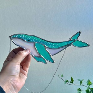 May include: A teal blue stained glass whale with white polka dots hanging from a silver chain. The whale has a white belly and a light blue tail.