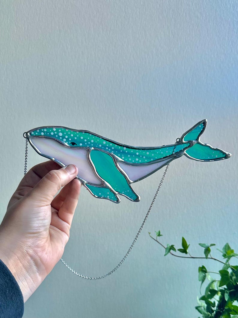 May include: A teal blue stained glass whale with white polka dots hanging from a silver chain. The whale has a white belly and a light blue tail.