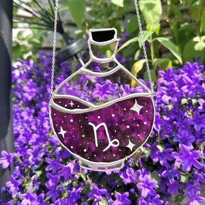 May include: A stained glass potion bottle with a purple liquid and white stars. The bottle has a silver chain for hanging and features the Capricorn zodiac symbol. The background includes purple flowers and green foliage.