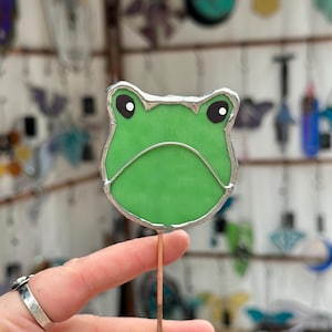 May include: A green stained glass frog with black eyes and a silver border. The frog is mounted on a copper rod. Other stained glass art pieces are visible in the background.