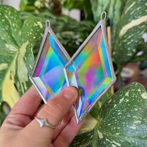 May include: Two diamond-shaped stained glass suncatchers featuring iridescent, multi-colored surfaces that shift between blue, pink, and yellow hues. Each piece is framed in a thin, silver-toned metal border and includes a small loop at the top.