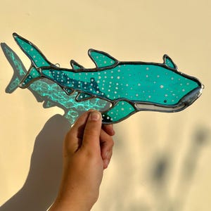 May include: A stained glass whale shark in shades of teal and white, with a silver-colored border. The shark has a spotted pattern and is held up against a light-colored background. The shark's fins and tail are visible.