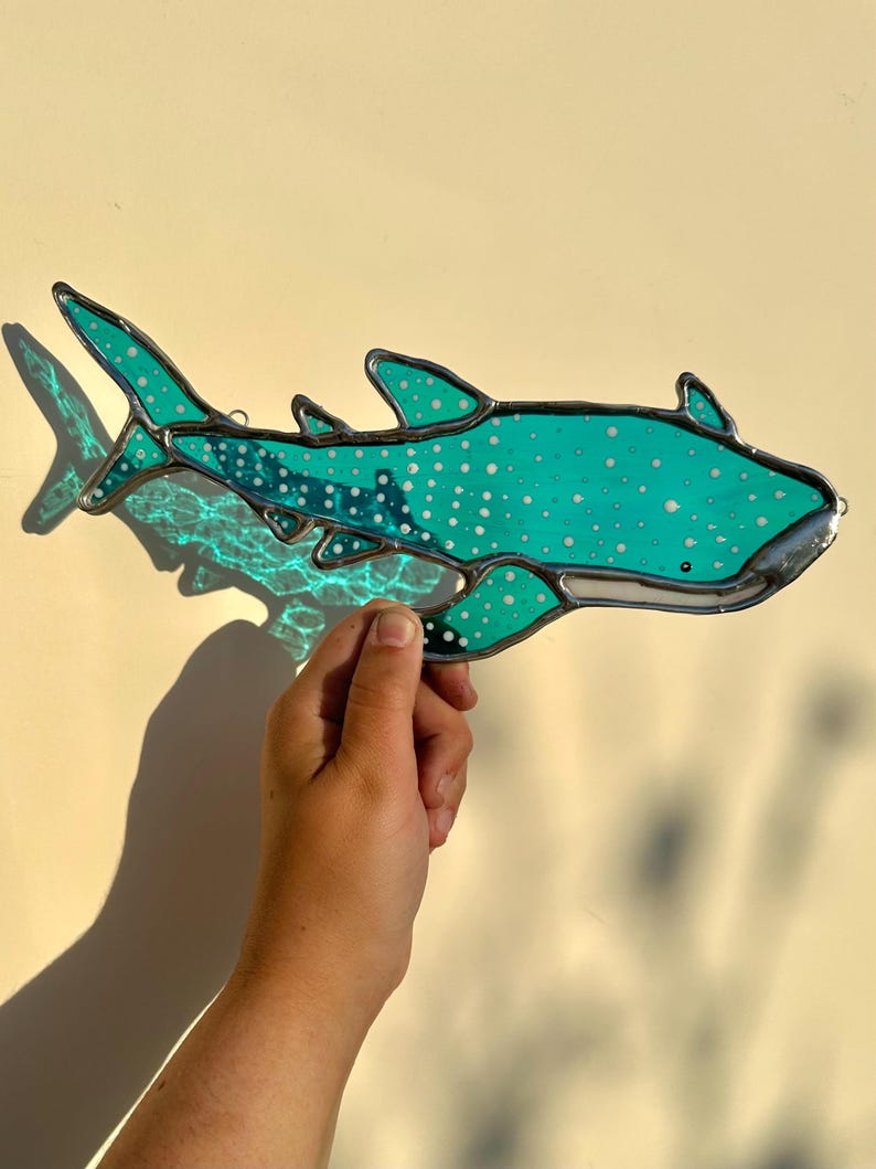 May include: A stained glass whale shark in shades of teal and white, with a silver-colored border. The shark has a spotted pattern and is held up against a light-colored background. The shark's fins and tail are visible.