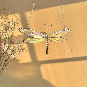 May include: A stained glass dragonfly with iridescent wings hanging from a silver chain. The dragonfly is suspended against a light beige background.