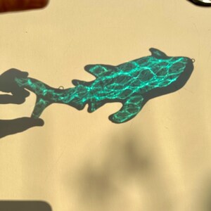 May include: A turquoise stained glass shark ornament with a textured surface. The shark is casting a shadow on a light beige surface. The ornament has a small loop for hanging.