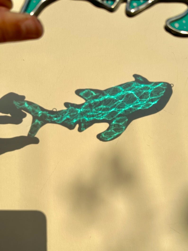May include: A turquoise stained glass shark ornament with a textured surface. The shark is casting a shadow on a light beige surface. The ornament has a small loop for hanging.