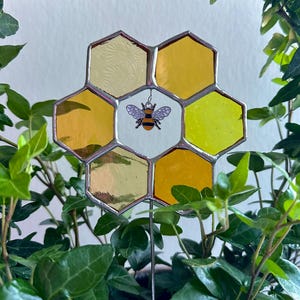 May include: A stained glass honeycomb with a bee in the centre. The honeycomb is made of yellow and gold glass and is surrounded by green leaves.