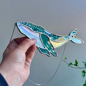 May include: A stained glass whale hanging from a silver chain. The whale is green, blue, and iridescent with white dots.
