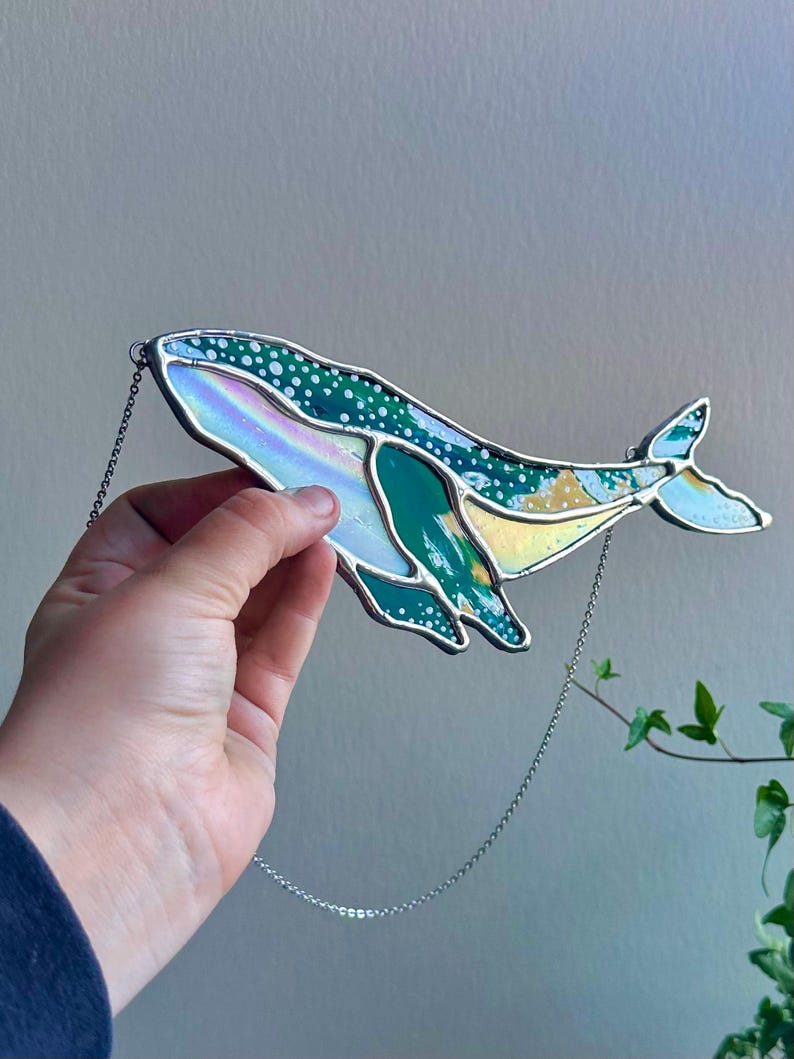 May include: A stained glass whale hanging from a silver chain. The whale is green, blue, and iridescent with white dots.