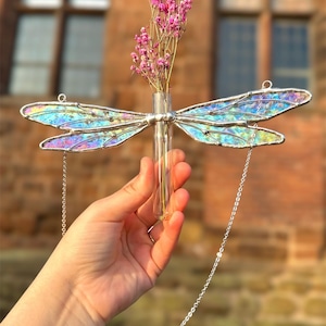 May include: A stained glass dragonfly with iridescent wings, holding a glass tube vase with pink flowers. The dragonfly is hanging from a silver chain.